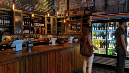 A cozy bar with wooden shelves filled with bottles, a counter, and a person browsing a drink fridge.