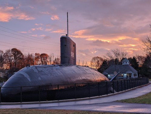 A submarine on display near a fence, with a colorful sunset and trees in the background.
