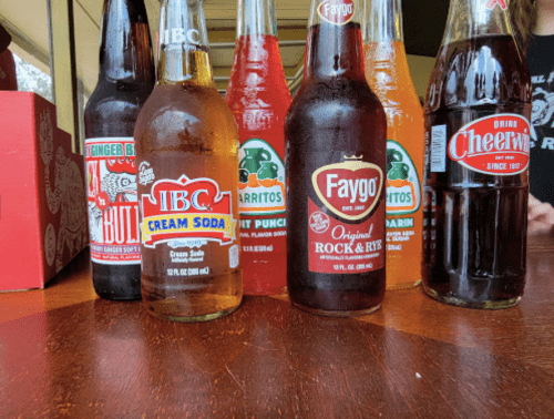 A variety of colorful soda bottles on a wooden table, including ginger beer, cream soda, and fruit punch.