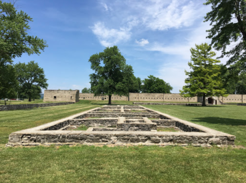 A grassy area with stone foundations and trees, set against a blue sky and historic stone walls in the background.
