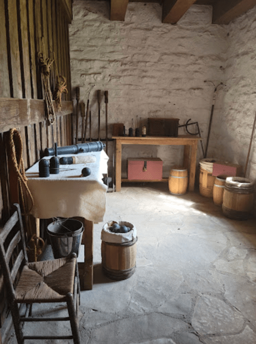 A rustic room with a wooden table, barrels, tools, and a chair, featuring stone walls and a simple, historical ambiance.