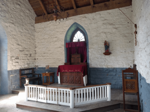 Interior of a rustic church featuring a wooden altar, stone walls, and a statue of a figure on the wall.