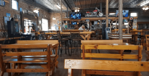 Interior of a rustic restaurant with wooden tables and chairs, bar area, and vintage decor, featuring TVs and a motorcycle.