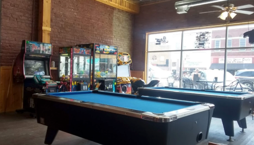 A spacious game room featuring a blue pool table, arcade machines, and large windows with a view of the street outside.