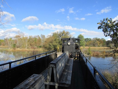 A vintage train car on a bridge overlooking a calm river, surrounded by trees and a blue sky with clouds.