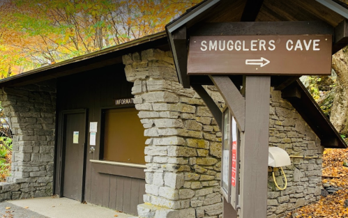 A stone building with a sign pointing to "Smugglers Cave" and an information window. Autumn leaves in the background.