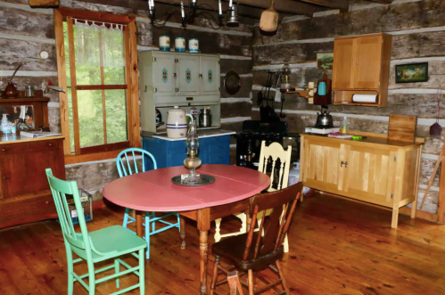 A rustic kitchen with a wooden table, colorful chairs, vintage appliances, and log cabin walls.
