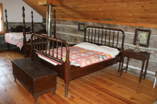 A rustic bedroom with wooden floors, two beds with quilts, a nightstand, and a vintage lamp under a wooden ceiling.
