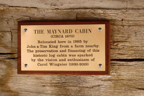 A plaque on a wooden wall reads about the Maynard Cabin, relocated in 1985, highlighting its preservation and history.