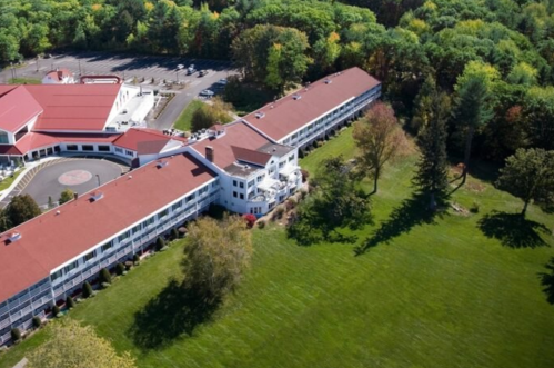 Aerial view of a hotel complex surrounded by green trees and a parking lot, featuring red roofs and landscaped grounds.