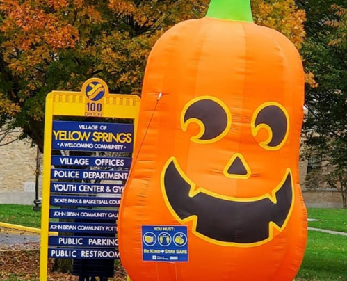 A large inflatable pumpkin stands next to a sign for the Village of Yellow Springs, surrounded by autumn foliage.