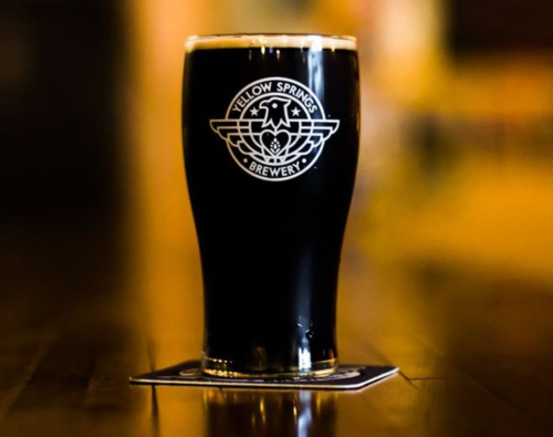 A dark beer in a glass with the logo of Fellow Springs Brewery on a wooden table.