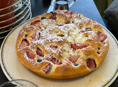 A round fruit cake topped with powdered sugar, featuring strawberries and blueberries, on a white plate.