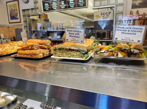 A display of sandwiches and a pizza in a deli, with signs for pesto chicken salad and steak sandwiches.