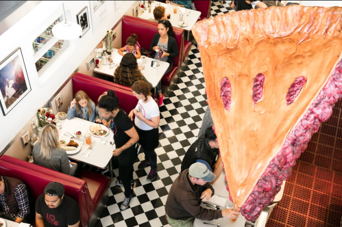 A busy diner scene with people eating at booths, featuring a large slice of pie in the foreground.