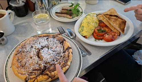 A plate with a large chocolate chip pancake dusted with powdered sugar, alongside an omelet, toast, and sliced tomatoes.