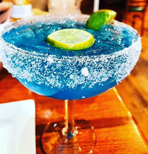 A blue cocktail in a salt-rimmed glass, garnished with lime slices, sits on a wooden table.