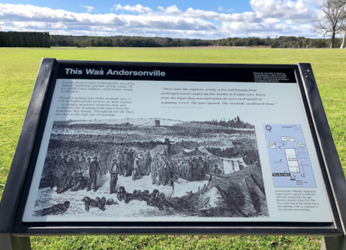 Sign about Andersonville, featuring historical information and an illustration of the prison camp. Green field in background.