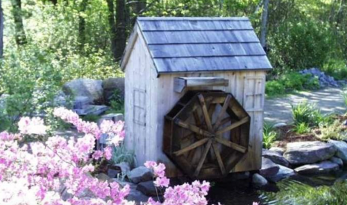 A small wooden watermill with a wheel, surrounded by blooming flowers and greenery by a peaceful pond.