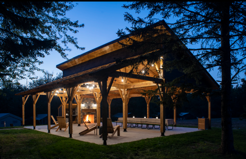 A wooden pavilion with lights, seating, and a fireplace, set in a serene outdoor environment at dusk.