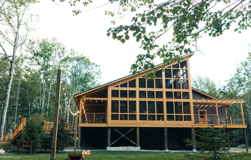 A modern wooden cabin with large windows, surrounded by trees and greenery, featuring a deck and outdoor seating.