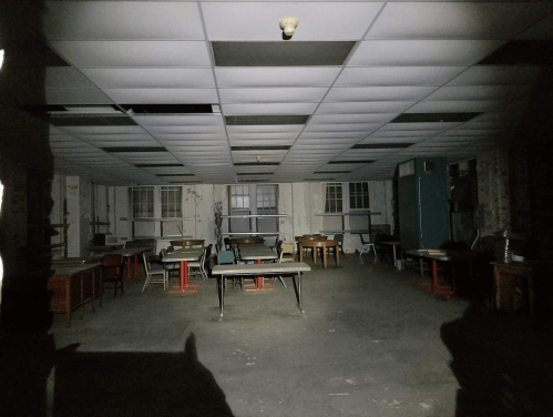 Dimly lit room with empty tables and chairs, featuring peeling walls and a ceiling with missing tiles.