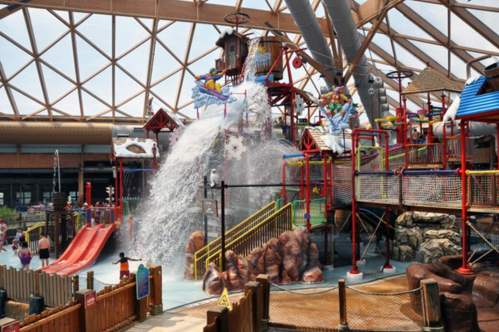 A vibrant indoor water park featuring slides, water features, and play structures under a large glass roof.