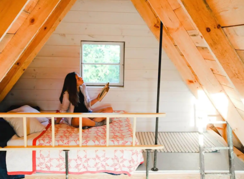 A woman sits on a bed in a cozy attic, reading a book by a window with wooden beams overhead.