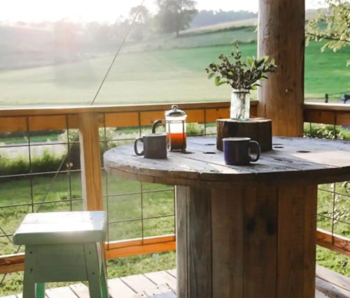 A rustic wooden table on a porch with two mugs, a French press, and a vase, overlooking a green landscape.