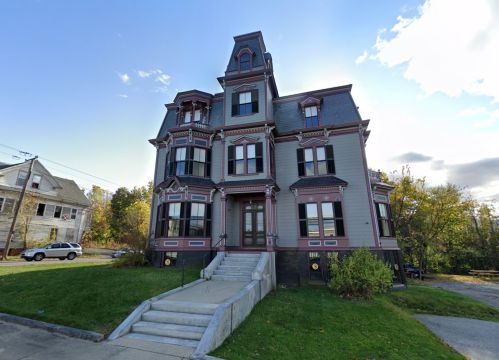 A large, ornate Victorian-style house with a steep roof and decorative trim, surrounded by greenery and a clear sky.