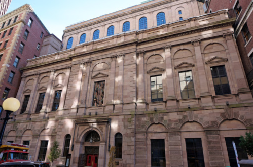 Historic stone building with large windows and decorative architectural details, set in an urban environment.