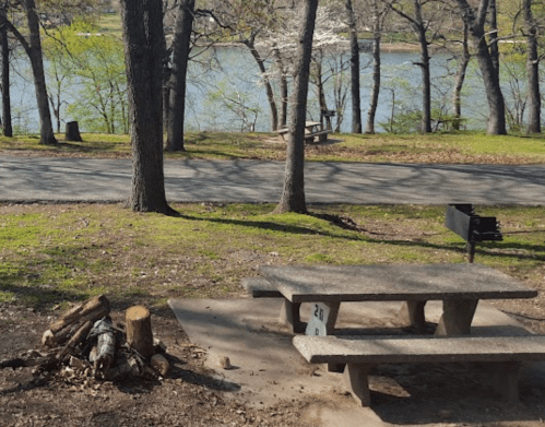 A picnic area with a stone table and fire pit, surrounded by trees near a calm lake.