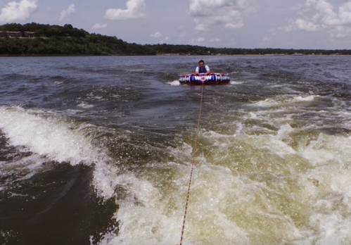 A person on a colorful inflatable tube being towed on a lake, with waves and a scenic shoreline in the background.