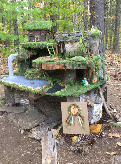 A whimsical fairy house made of wood and moss, surrounded by pinecones and autumn leaves in a forest setting.