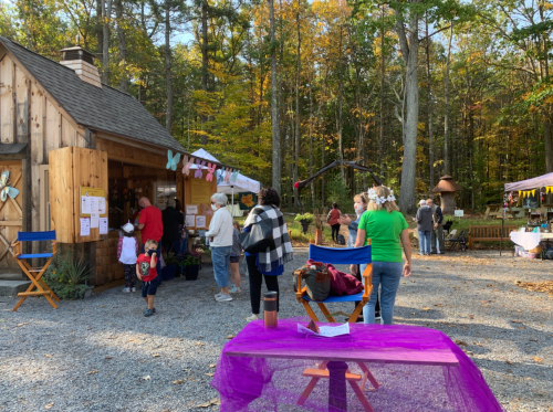 People gather at a rustic outdoor market surrounded by trees, with colorful decorations and stalls in the background.