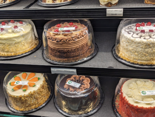 A display of various decorated cakes in clear containers, including carrot cake, chocolate, and vanilla options.