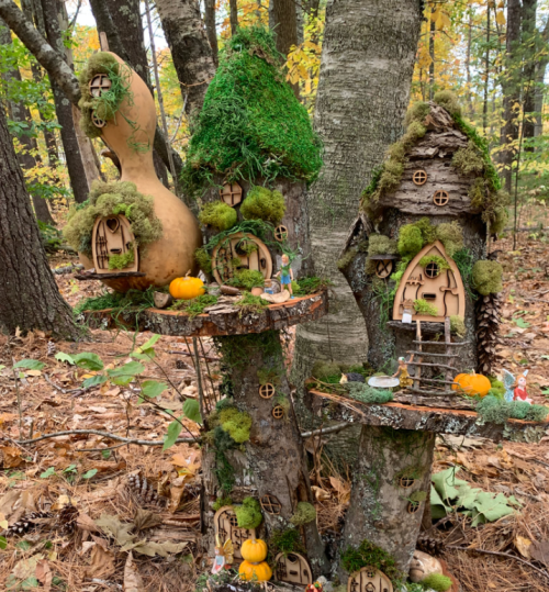 Whimsical fairy houses made of wood and moss, nestled among trees with autumn leaves and small pumpkins.