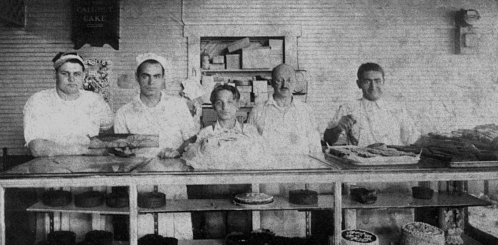 Black and white photo of five bakers in a shop, standing behind a counter filled with various cakes and pastries.