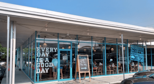 A modern BBQ restaurant with large windows, featuring a sign that says "Every day is a good day" and outdoor seating.