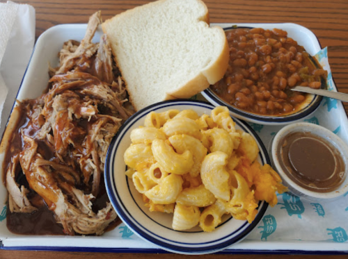 A tray with pulled pork, macaroni and cheese, baked beans, a slice of bread, and a small cup of sauce.