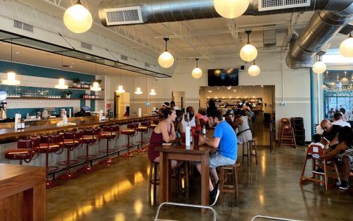 A modern restaurant interior with bar seating, patrons dining, and pendant lights hanging from the ceiling.
