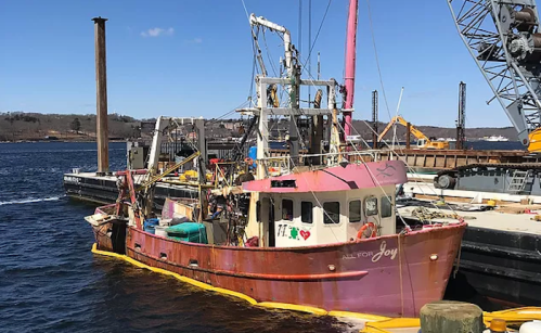 A colorful fishing boat named "All for Joy" docked at a harbor, surrounded by water and other vessels.