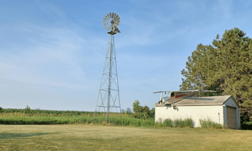 A windmill stands beside a small shed with an airplane parked nearby, surrounded by tall grass and trees.
