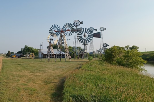 A row of vintage windmills stands near a grassy area and a calm waterway under a clear sky.