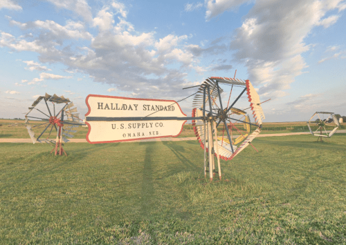 Sign for Halladay Standard U.S. Supply Co. with decorative windmills in a grassy field under a cloudy sky.