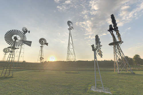 A collection of vintage windmills silhouetted against a sunset in a grassy field.