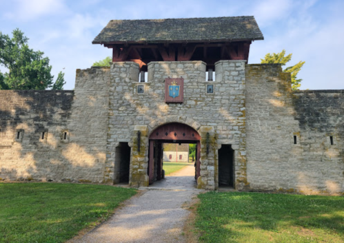 A stone castle gate with a wooden roof, flanked by walls and greenery, leading to an open courtyard.