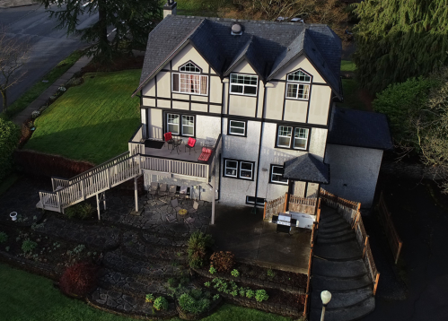 Aerial view of a large, two-story house with a deck, surrounded by green grass and trees.