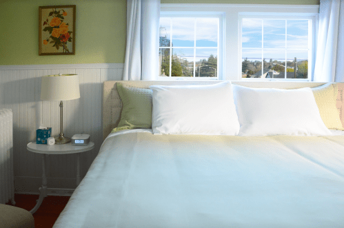 A cozy bedroom with a neatly made bed, a bedside table, and large windows letting in natural light.