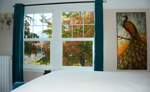 A cozy room with a bed, two windows showing colorful autumn trees, and a peacock painting on the wall.